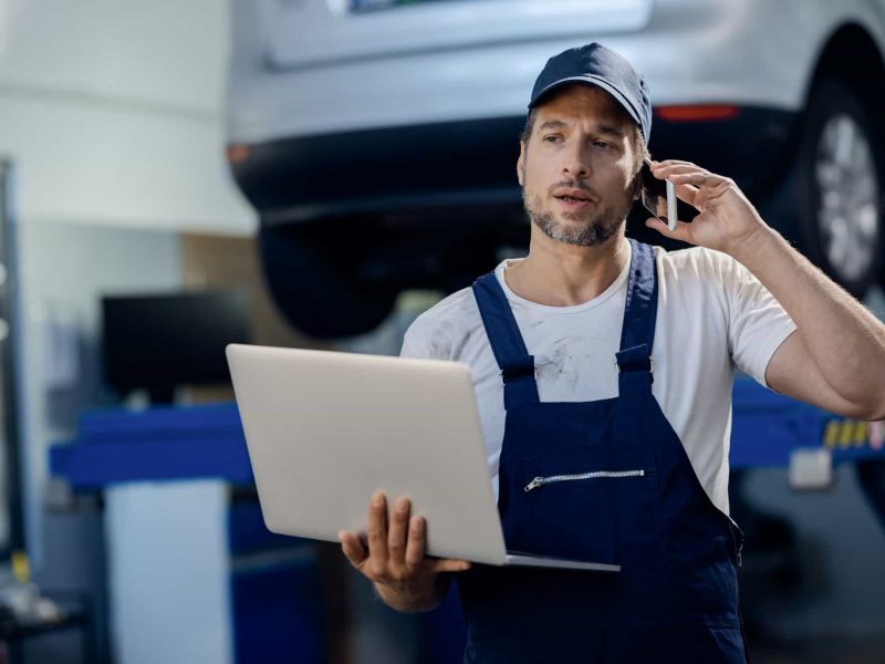 auto-repairman-talking-on-the-phone-while-using-laptop-in-a-workshop-.jpg
